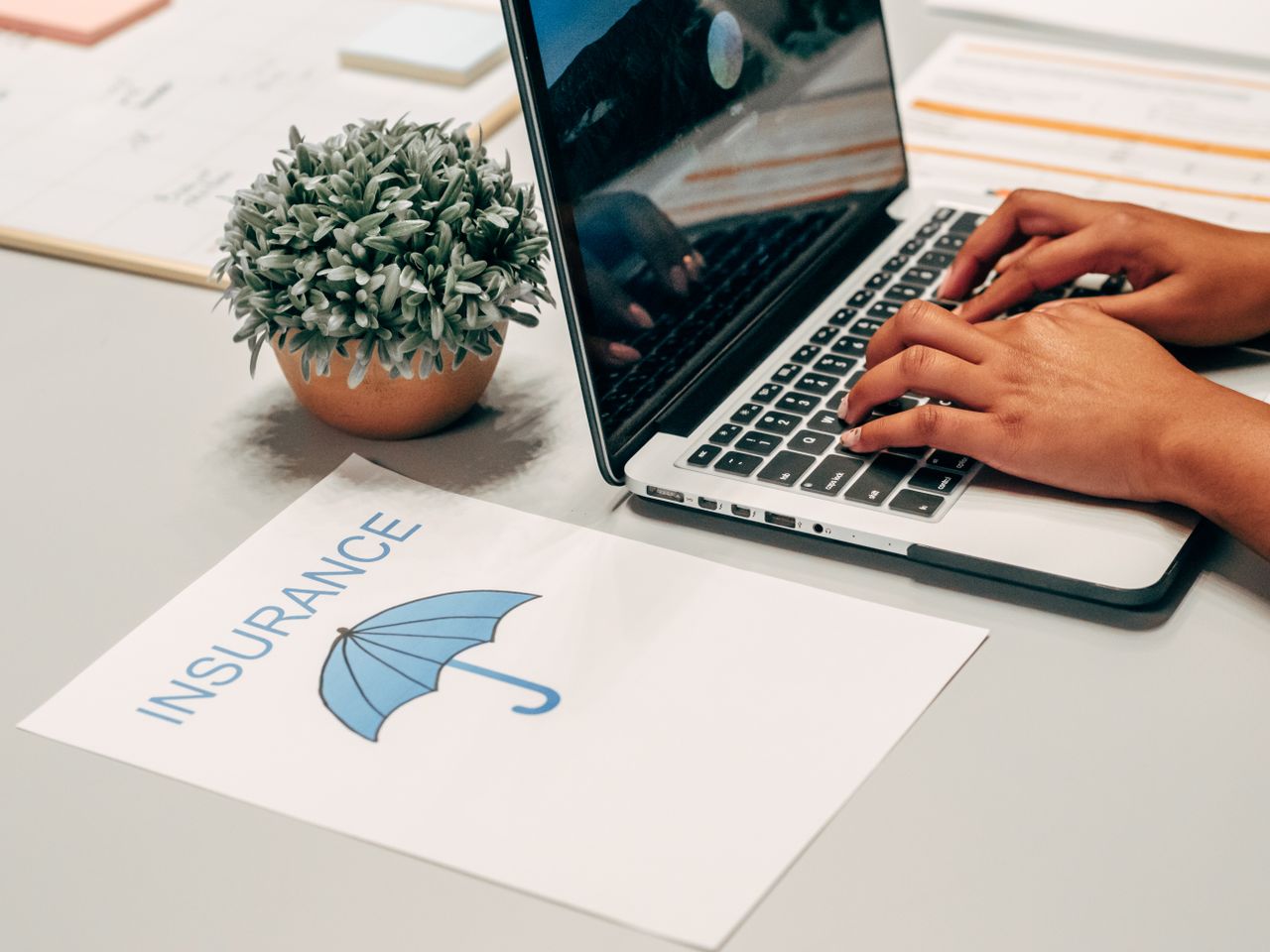 about-us Hands typing on a laptop at a desk with an insurance paper and plant, suggesting a work environment.